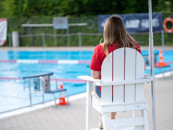 Rettungsschwimmerin auf dem Aufsichtsstuhl am Beckenrand – Freibad Dellwig Eine Rettungsschwimmerin in rotem T-Shirt sitzt auf einem weißen Aufsichtsstuhl unter einem roten Sonnenschirm und blickt über das blaue Schwimmerbecken des Freibads. Im Hintergrund Startblöcke, Leinen und ein Rettungsring.